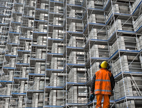 Construction worker wearing safety gear inspecting metal scaffolding at a large building site.