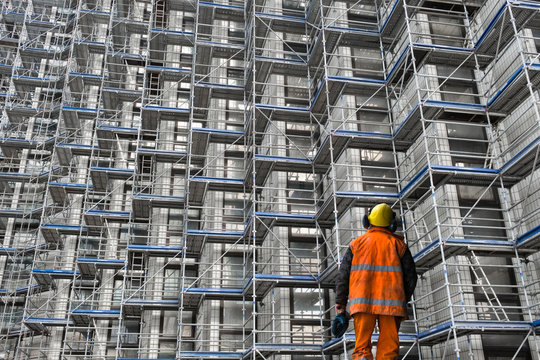 Construction worker wearing safety gear inspecting metal scaffolding at a large building site.