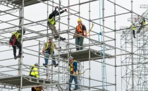 Construction workers wearing safety harnesses and helmets assembling metal scaffolding at a building site.