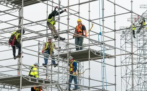 Construction workers wearing safety harnesses and helmets assembling metal scaffolding at a building site.