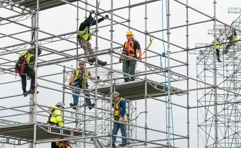 Construction workers wearing safety harnesses and helmets assembling metal scaffolding at a building site.