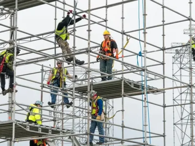 Construction workers wearing safety harnesses and helmets assembling metal scaffolding at a building site.
